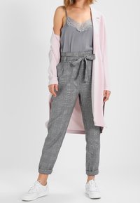 Cappotto lungo rosa chiaro sopra un top in pizzo grigio, abbinato a pantaloni a quadri grigi con vita a cravatta e sneakers bianche.