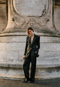 Zwart gestreept pak met diepe V-hals en lange mouwen, met een nauwsluitende pasvorm en een gladde textuur. Accessoires omvatten grote oorbellen.