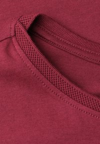 Gros plan d'un tissu bordeaux montrant une couture côtelée et texturée au niveau de l'encolure d'un vêtement en coton.