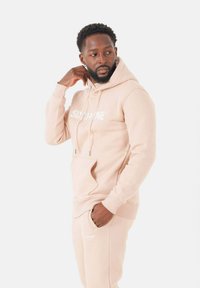 Hoodie et joggers beige clair en tissu doux. Comprend une poche frontale et un logo blanc sur la poitrine, avec des accents de cordon de serrage.