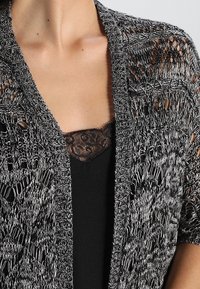 Svart och vit spetskofta med öppen fram, texturerad stickad design och korta ärmar, lager på en svart spets-cami-top.