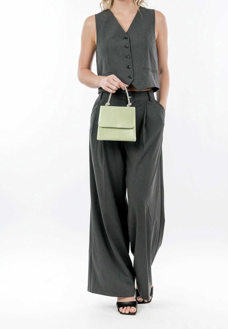 Femme portant un gilet boutonné gris foncé et un pantalon à jambes larges, tenant un sac à main vert clair, debout avec une main dans la poche.