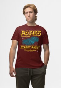 Burgundské bavlněné tričko s modrou grafikou závodního auta a tučným žlutým textem "PARIS", doplněné o další detaily s tématikou závodů.