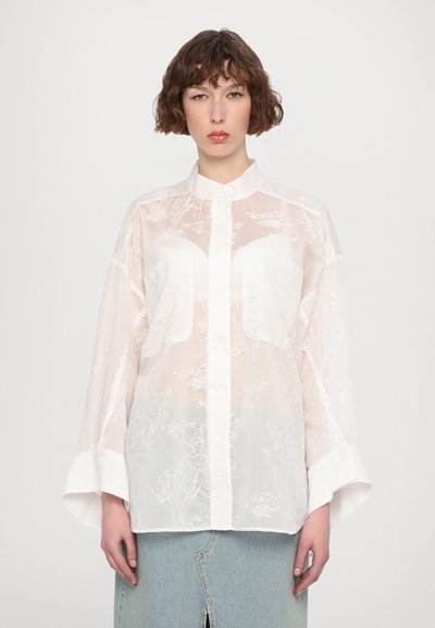 Blouse blanche en voile avec broderies florales, col haut, devant à boutons et manches larges. Conçue en tissu léger avec une texture subtile.