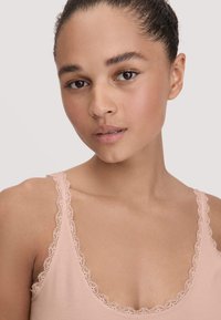 Close-up of a woman with tied-back hair wearing a light pink camisole with lace trim, facing forward with a neutral expression.