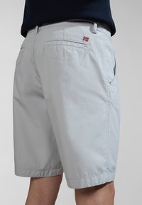 Lysgrå shorts lavet af åndbart stof, med sidelommer og en lille rød og blå flagdetalje på baglommen.