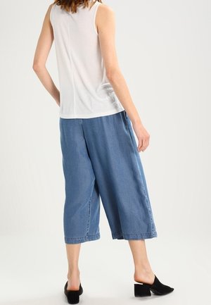 Weißes ärmelloses Oberteil kombiniert mit hellblauen, weit geschnittenen Denim-Hosen in mittlerer Wadenlänge. Schwarze Schuhe mit Blockabsatz runden das Outfit ab.