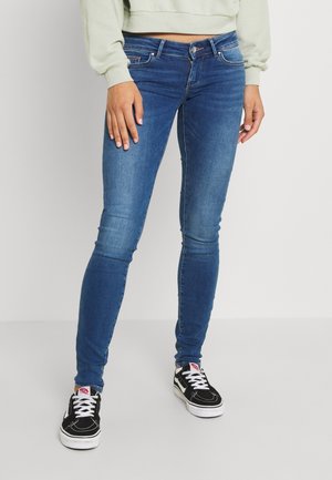 Person trägt blaue Skinny Jeans, schwarz-weiße Sneakers und ein hellfarbenes Cropped Sweatshirt und steht vor einem einfarbigen Hintergrund.