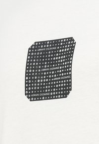 Quadrado preto com cantos arredondados preenchido com fileiras de pontos brancos e o texto repetido "ARMANI EXCHANGE" em tecido branco.