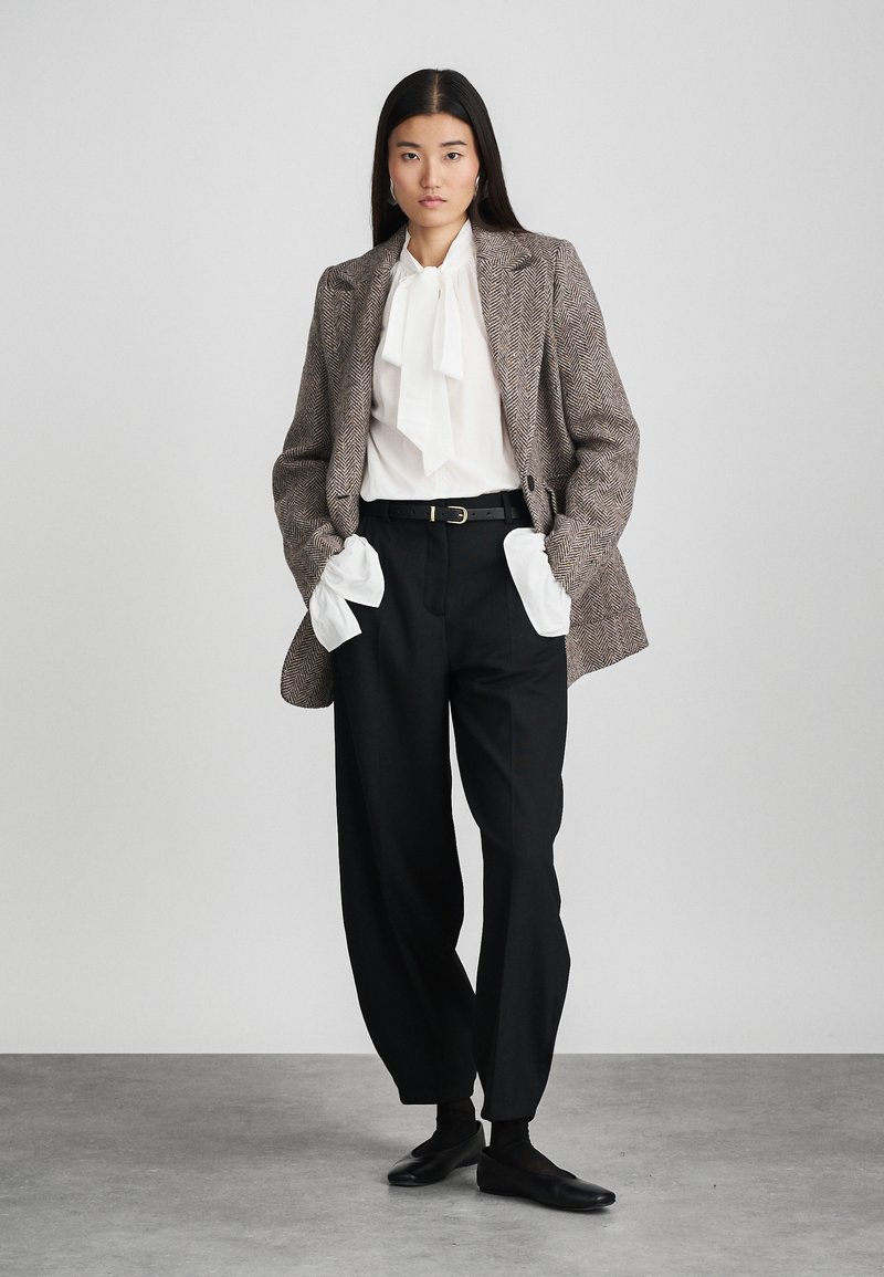 Blazer marrone a spina di pesce con spalle strutturate, camicia bianca con fiocco, pantaloni neri a vita alta e mocassini neri.