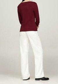 Maglietta a maniche lunghe color bordeaux con scollo a barca e zip sul retro, abbinata a pantaloni bianchi a gamba larga e con vita alta e scarpe nere.