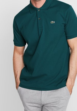 Ciemnozielona koszulka polo z teksturowanej tkaniny, z zapięciem na dwa guziki i małym haftowanym logo po lewej stronie klatki piersiowej. Połączona z szarymi spodniami.