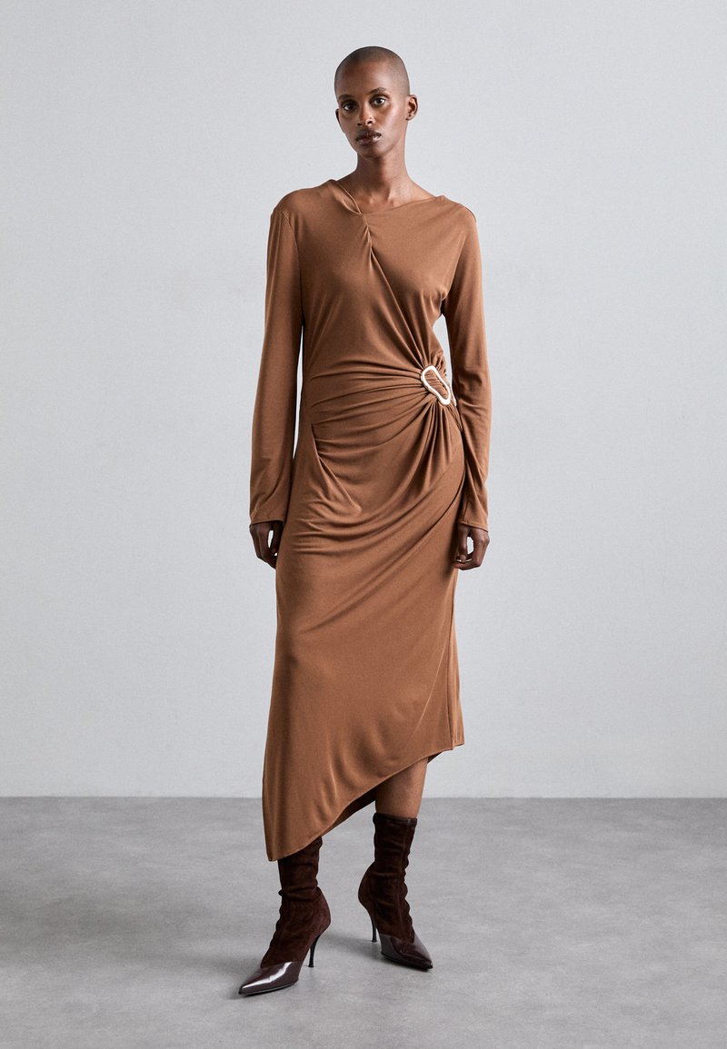 Vestido marrón de manga larga con detalle de nudo en la cintura, dobladillo asimétrico y textura suave, combinado con botas de tobillo marrón oscuro.