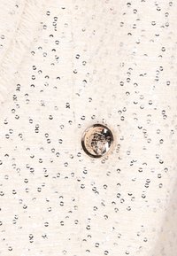 Krémová látka se stříbrnými flitry a lesklým zlatým knoflíkem s dekorativním emblémem. Texturovaný povrch s detaily třásní.
