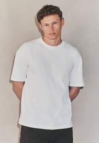 Vit bomullst-shirt med rund hals och korta ärmar, med en avslappnad passform och slät textur. Bärs med mörka byxor.