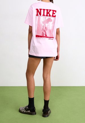 Person trägt ein hellrosa Nike-T-Shirt mit roter Grafik und Text, schwarze Shorts, schwarze Socken und zebra gemusterte Schuhe und steht auf grünem Boden.