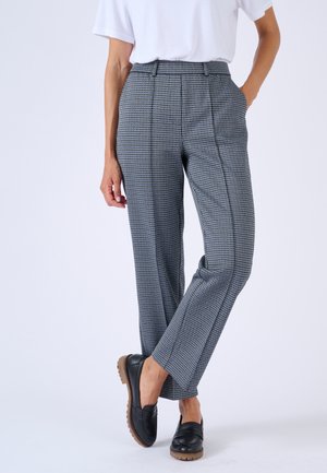 Femme portant un pantalon droit gris et bleu à carreaux avec des plis à l'avant, une chemise blanche à manches courtes, des mocassins noirs, debout avec une main dans la poche.