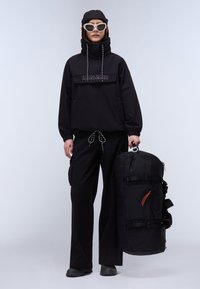 Svart vanntett jakke med høy krage, elastiske mansjetter og logodetalj. Svart cargo-bukse og svart duffelbag med orangemerk.