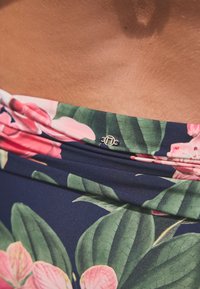 Maudymosi kostiumėlio viršus su gėlių raštu, kuriame pavaizduotos rožinės gėlės ir žali lapai tamsiai mėlyname fone, puoštas dekoratyvine metaline detalė.