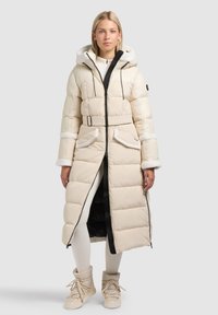 Lange beige donzen jas met capuchon, taille met riem en witte accenten. Heeft een gewatteerd ontwerp en zijzakken, gedragen over lichte broek en laarzen.