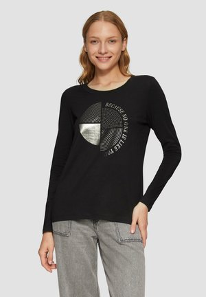 Schwarzes Langarmshirt mit einem kreisförmigen grafischen Design und Linien sowie einem silbernen Akzent, kombiniert mit hellgrauen hoch taillierten Jeans.