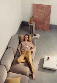 Grijze modulaire bank met stevige kussens, gestreepte lange mouwen top en gele broek. Lichte salontafel met een keramisch kopje en vaas.