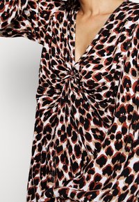 Kobieta ubrana w sukienkę z długim rękawem w leopardzi wzór, z przekręconym węzełkiem na klatce piersiowej i dekoltem w kształcie litery V.