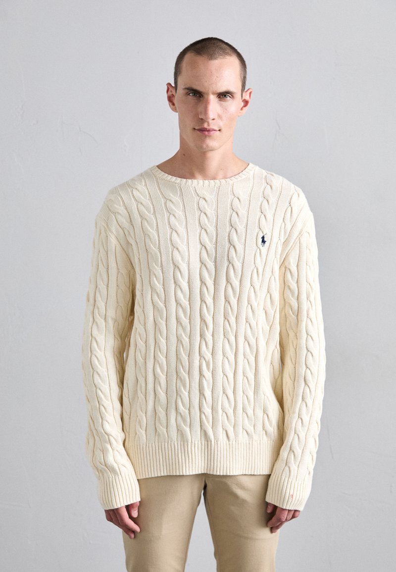 Cream cable-knit sweater with ribbed cuffs and hem, featuring a small blue logo on the left chest. Worn with light beige pants.