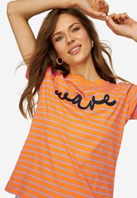 Camiseta de manga corta naranja con rayas moradas. Presenta un gráfico en cursiva negra de "ola" en la parte frontal. Material de algodón, ajuste relajado.