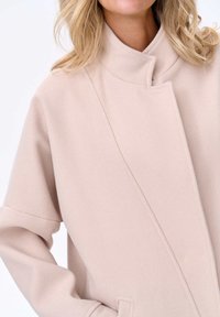 Beige coat gemaakt van zachte stof met een ontspannen pasvorm. Kenmerkt zich door een staande kraag en zijzakken. Strakke lijnen en minimalistisch ontwerp.