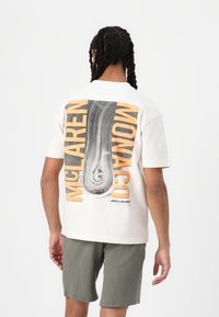 Lys beige bomulds t-shirt med et sort og orange grafisk tryk af en racerbane på ryggen, med teksten "MCLAREN MONACO" ovenover.