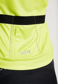 Top atlético amarillo fluorescente con cintura negra, que cuenta con una cremallera discreta y un logotipo reflectante en el dobladillo. Tejido suave y liviano.