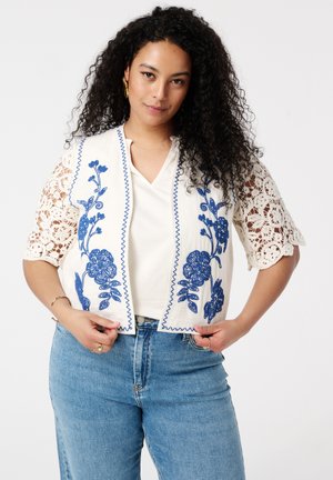Gilet blanc brodé avec des motifs floraux bleus, manches en dentelle festonnées et design en V, associé à un haut blanc uni et un jean en denim.