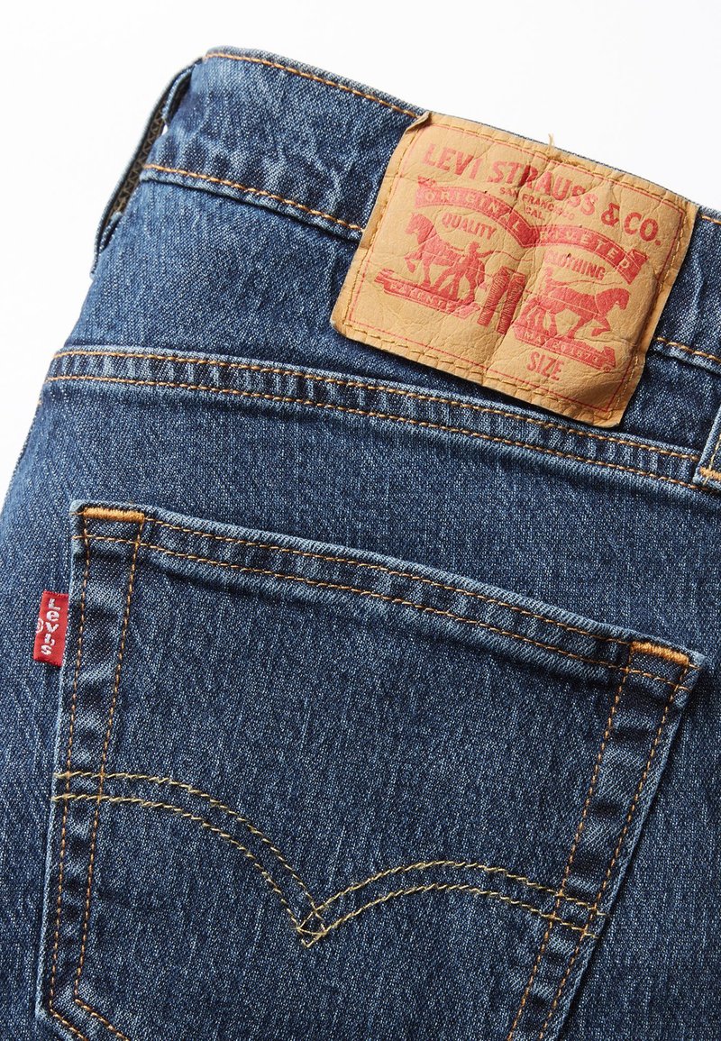 Jean en denim bleu avec une patch en cuir brun arborant le logo Levi Strauss & Co. Les coutures sont orange, avec une petite étiquette rouge sur la poche arrière.