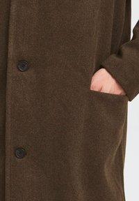 Brun ullblandingsfrakk med en myk tekstur, med knappelukking og sidelomme, som fanger stoffets varme og dybde.