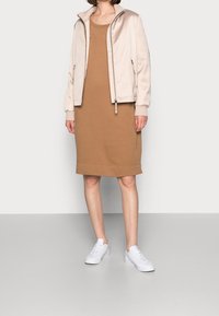 Beige bomberjacka med fickor med dragkedja över en brun ärmlös klänning. Vita sneakers fulländar outfiten. Släta texturer, moderna former.