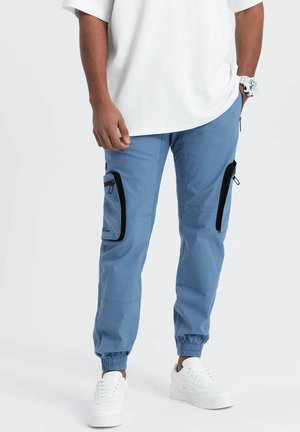 Hellblaue Cargo-Hosen mit schwarzen Reißverschlüssen und Taschen, elastischen Bündchen und einer lockeren Passform, kombiniert mit weißen Sneakers und einem lockeren weißen Hemd.