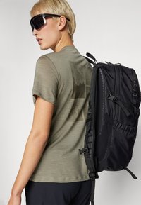 Alyvuogių spalvos marškinėliai su subtiliu grafinio dizaino akcentu, derinami su dideliu juodu kuprinė, turinčia reguliuojamus diržus ir tekstūrinius šoninius kišenes.