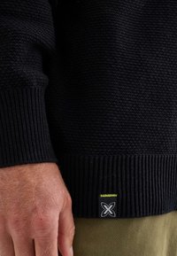 Detail eines schwarzen Strickpullovers, der den strukturierten Stoff, das gerippte Bündchen und dasLogoschild mit gelben Nähten zeigt. Eine Hand ruht auf dem Stoff.