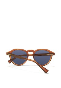 Kulaté sluneční brýle s průsvitným jantarovým rámem a modrými čočkami. Mají jedinečný šikmý most a logo Hawkers na stranicích.