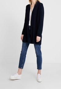 Granatowy dzianinowy kardigan z głębokim dekoltem w serek, prążkowaną fakturą, długimi rękawami i otwartym przodem, zestawiony z jasnoniebieskimi jeansami i białymi trampkami.