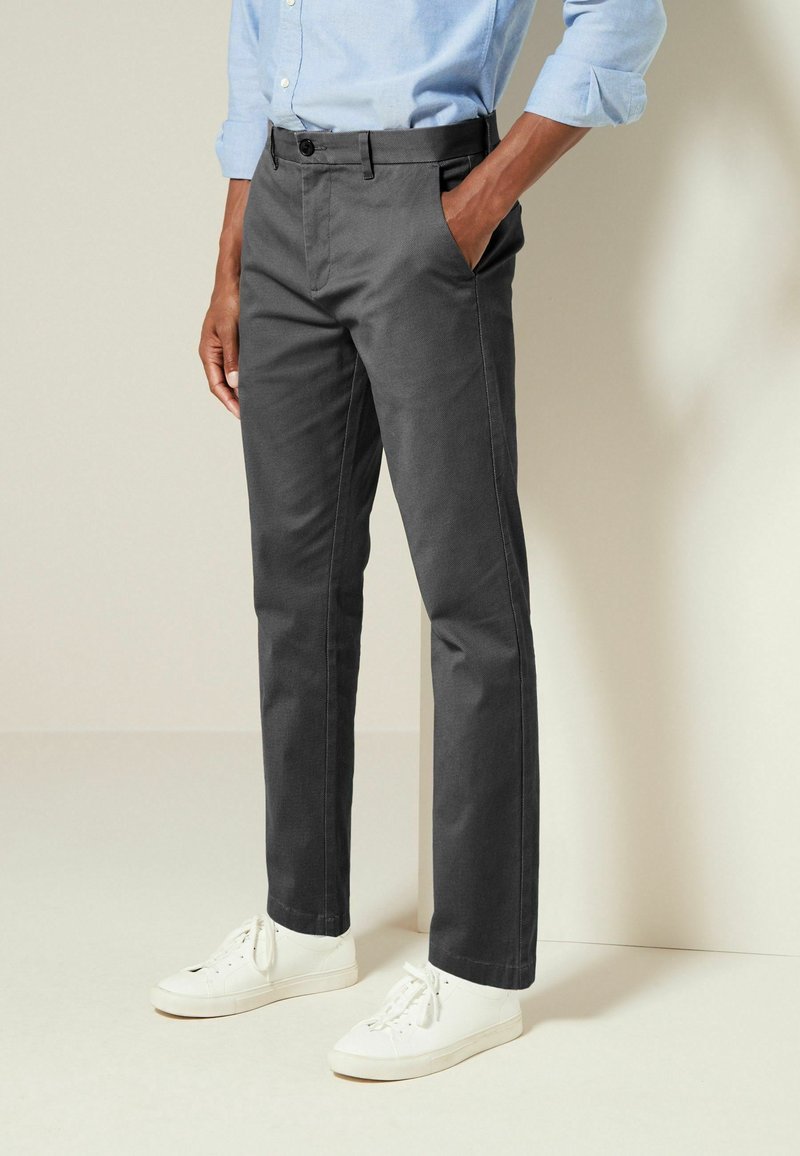 Homme portant un pantalon droit gris foncé, des baskets blanches et une chemise bleu clair à manches longues avec une main dans la poche.