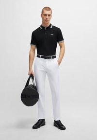 Polo noir à manches courtes avec des accents blancs, pantalon blanc, ceinture et chaussures noires ; tenant un grand sac de sport noir avec des poignées.