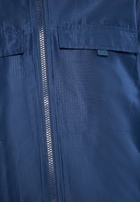 Tamsiai mėlyna striukė su tekstūruota paviršiaus, centrinė užtrauktuką ir dviem krūtinės kišenėmis su atvirtimu. Medžiaga atrodo lengva ir glotni.