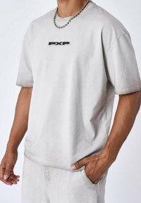 Camiseta de manga corta gris claro en tejido suave, con un logotipo rectangular negro "PXP" en el pecho y un corte relajado.