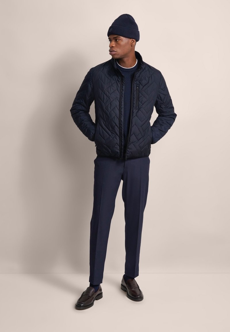 Navy gesteppte Jacke mit hohem Kragen, kombiniert mit einem navyfarbenen Strickpullover und maßgeschneiderten dunklen Hosen, ergänzt durch dunkle Slipper und eine Mütze.