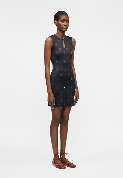 Czarna sukienka bez rękawów o dopasowanym kroju, z małymi czerwonymi wzorami wiśni, z dekoltem typu keyhole, noszona z czerwonymi płaskimi butami na sznurowadła.