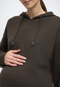 Σκούρα καφέ φούτερ με κουκούλα, άνετη εφαρμογή, κορδόνι ρύθμισης και μαλακή υφή, με ένα χέρι τοποθετημένο πάνω σε ορατή κοιλιά.