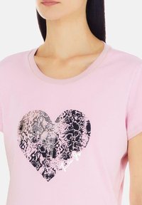 T-shirt rosa a maniche corte con un grande design a forma di cuore nero caratterizzato da dettagli testurizzati e una firma stampata bianca sotto il cuore.