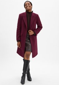 Vrouw die glimlacht in een bordeauxrode jas, een donkerbruine coltrui, een zwarte leren rok en zwarte kniehoge laarzen op een witte achtergrond.